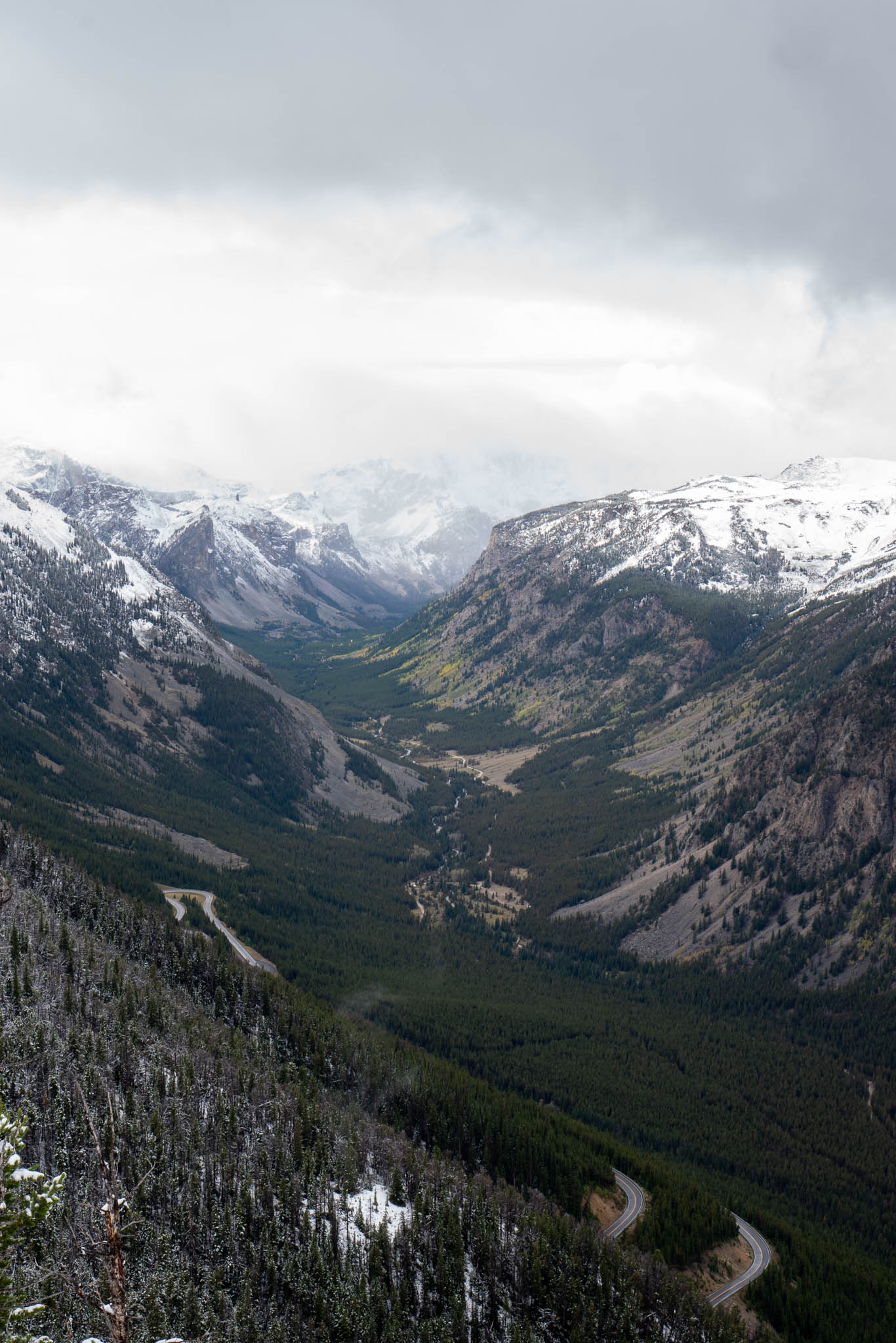 An alpine valley framed by snow-dusted peaks and drifting clouds, with a two-lane road winding through the forest below.