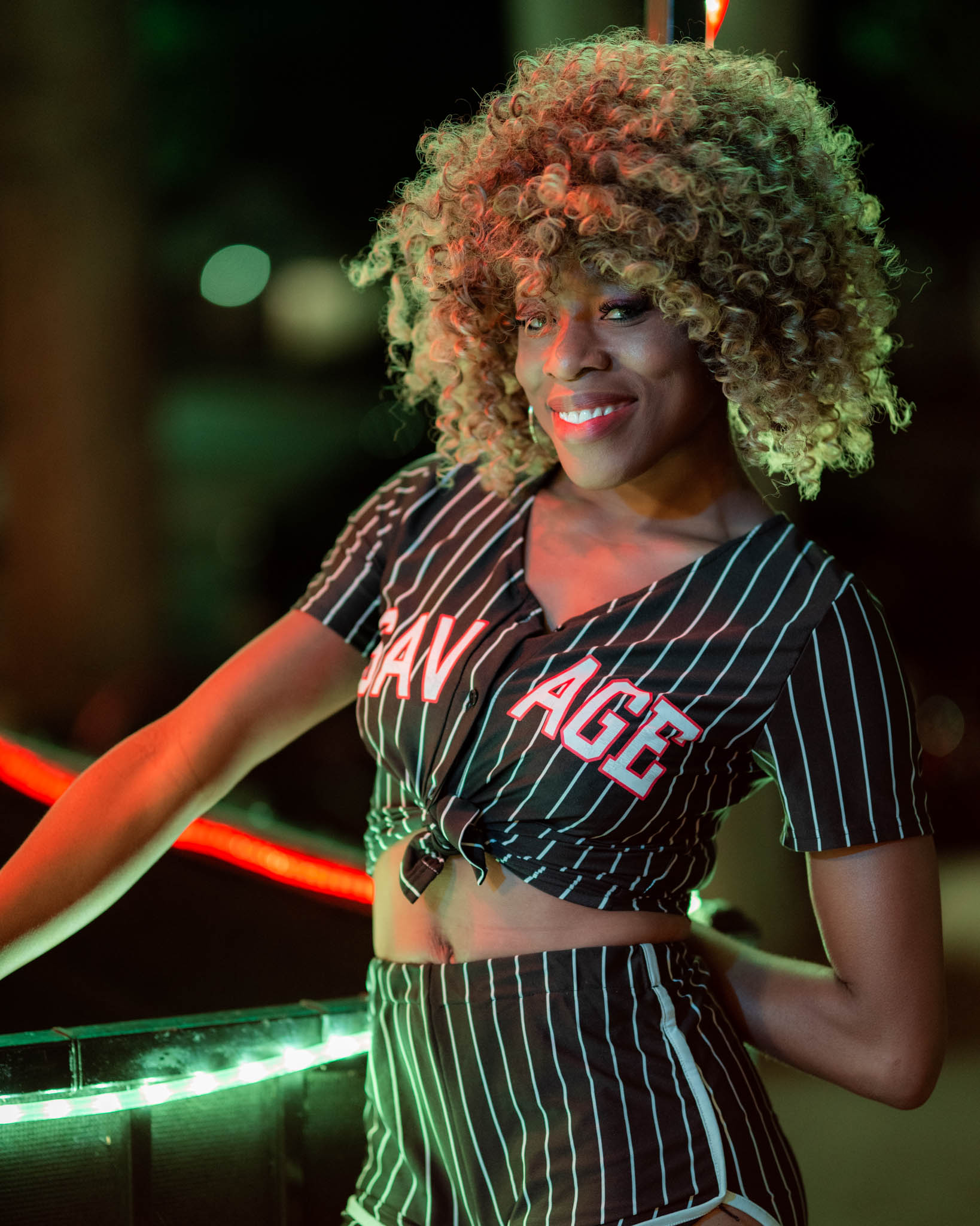 A woman in a pinstriped crop top and shorts smiles beside a glowing red neon light at night.