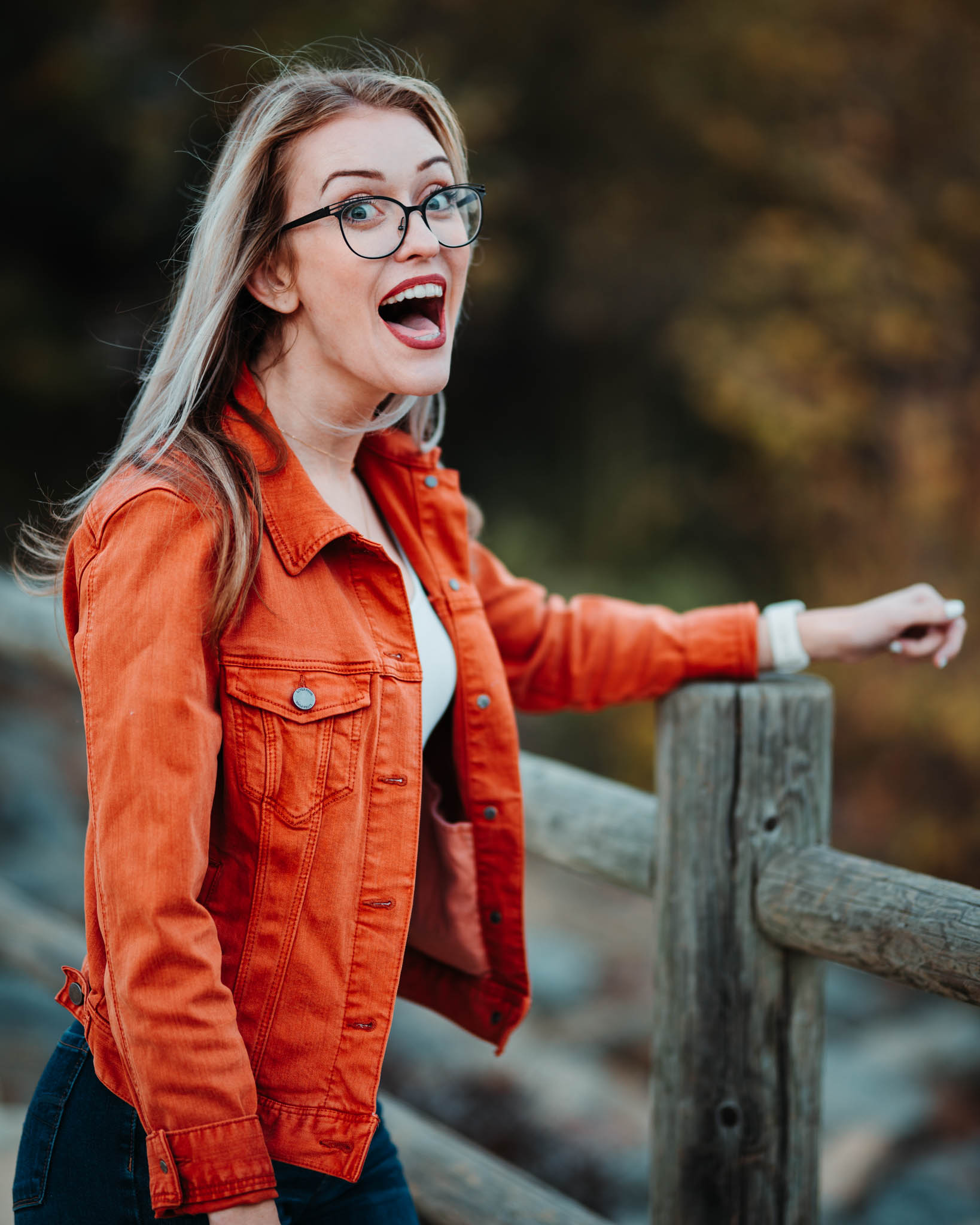 A woman in an orange denim jacket and glasses laughs while leaning on a wooden fence rail.
