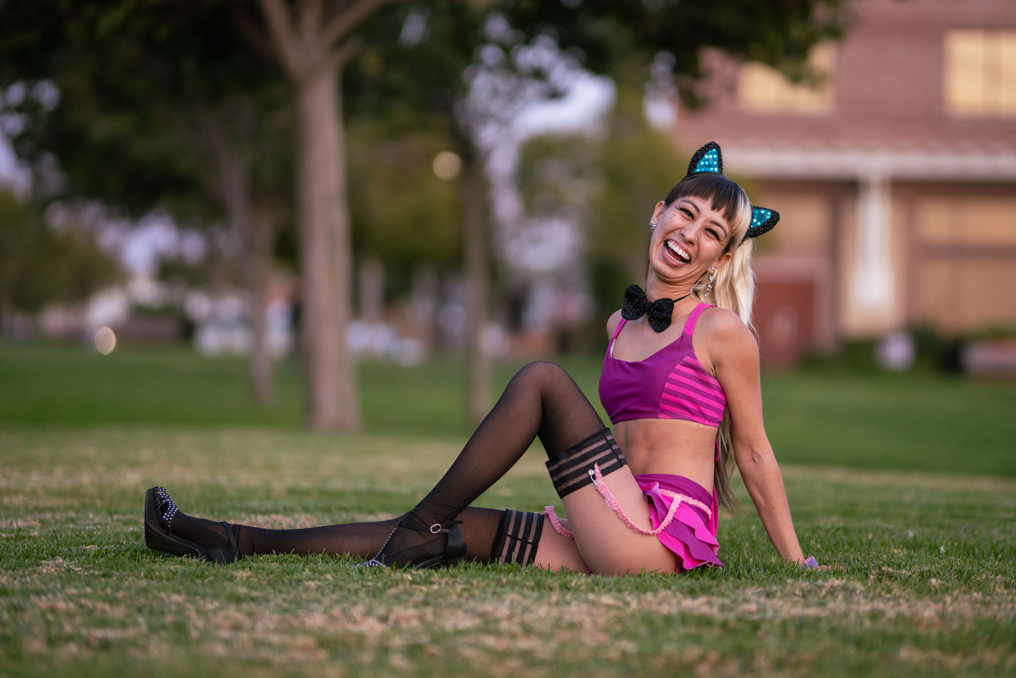 A woman in a pink cat-ear headband and striped bralette laughs while sitting on a lawn at dusk.