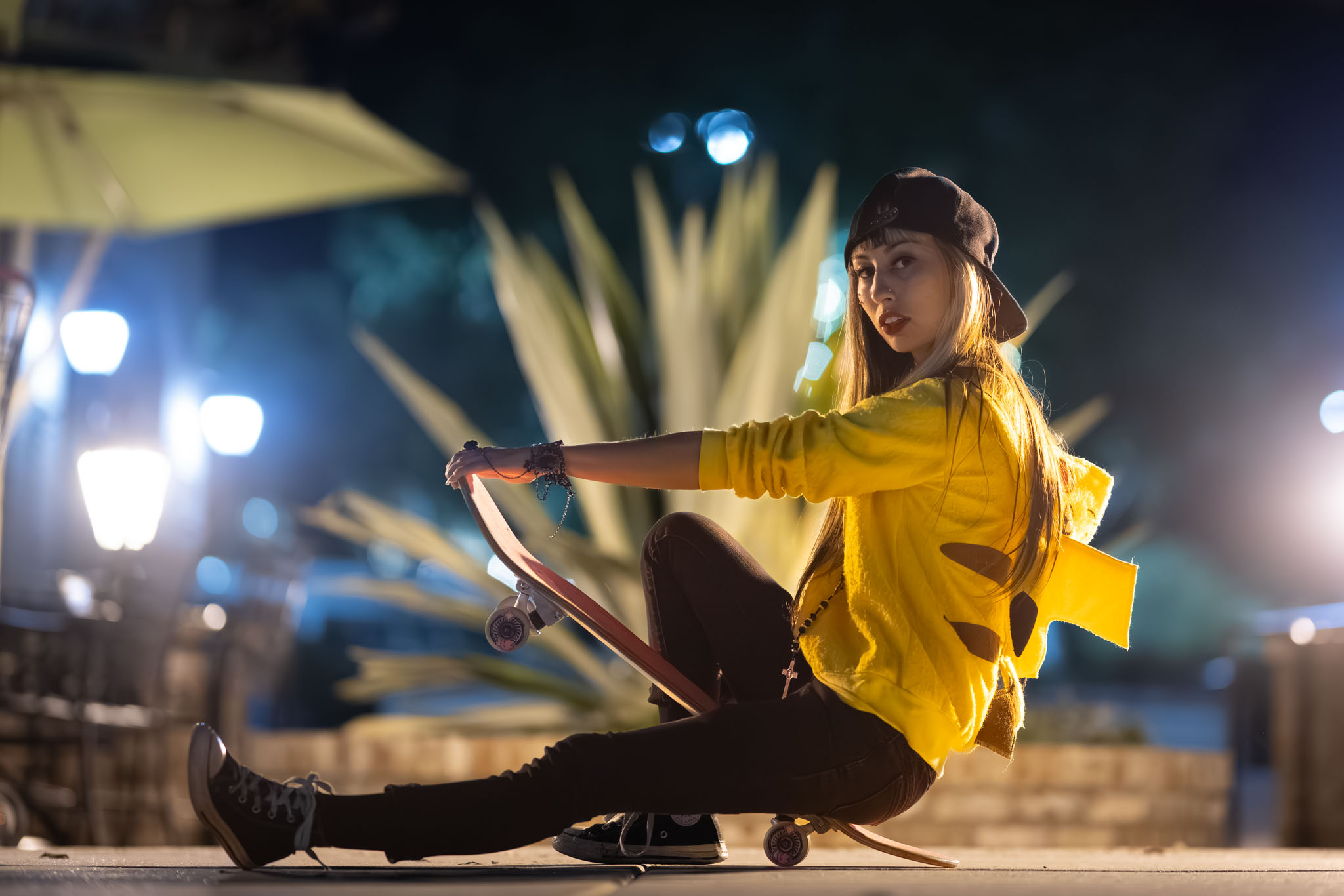 A woman in a yellow Pikachu hoodie sits on a skateboard at night, string lights glowing behind her.