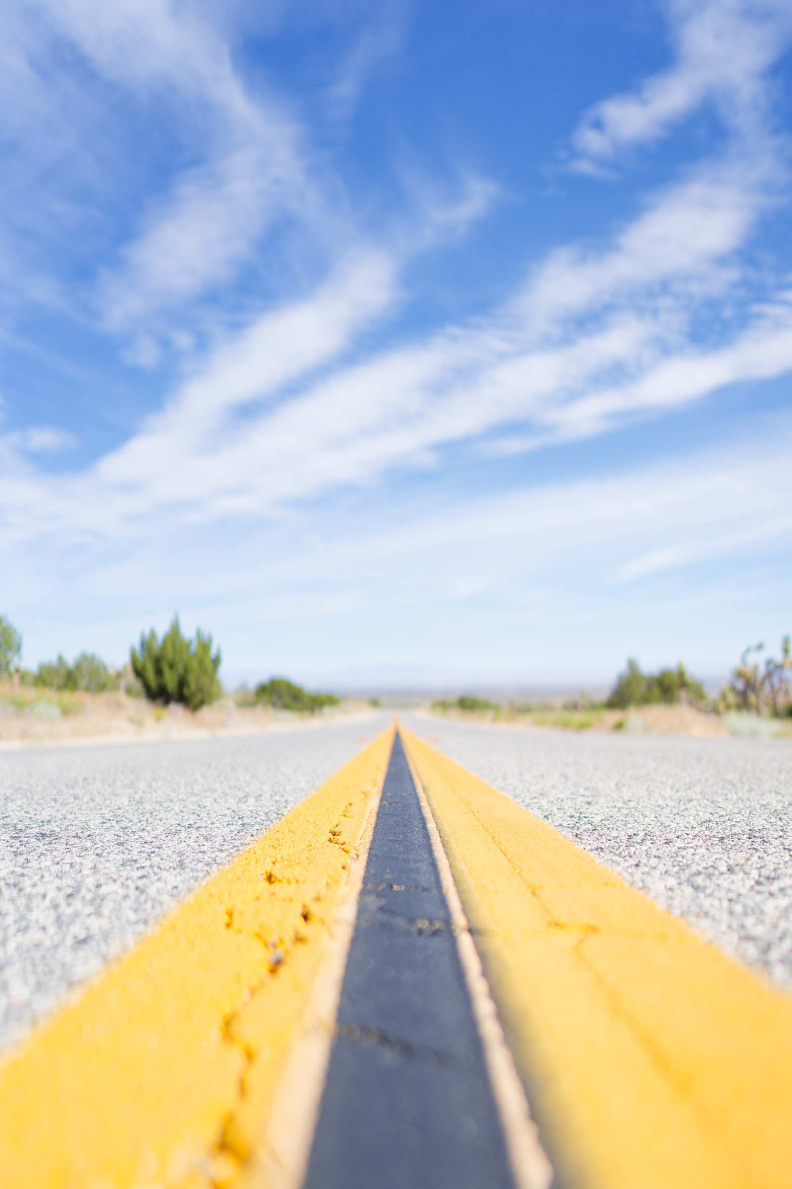 Low angle down a two-lane desert highway, the double yellow center line leading to a distant horizon under a wide blue sky.